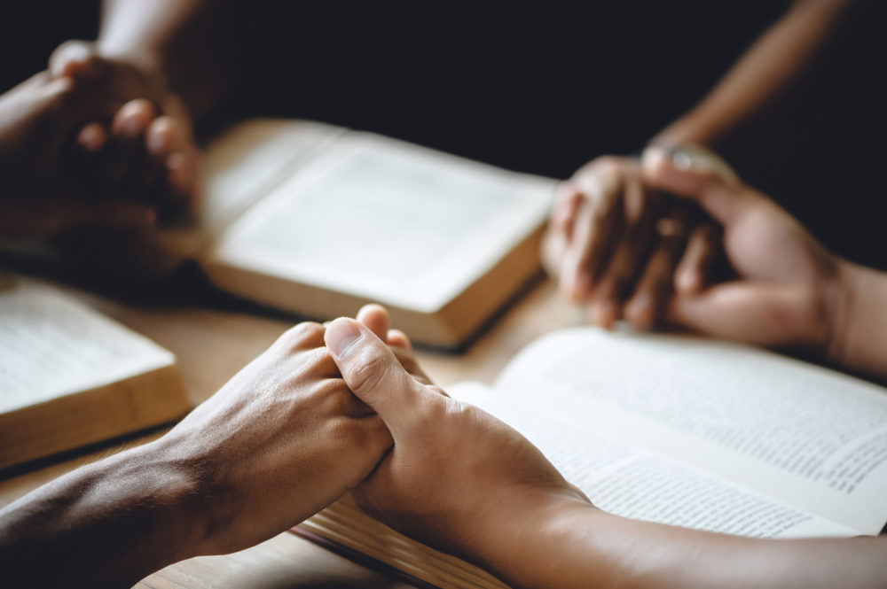 Christian group of people holding hands praying worship to believe and Bible on a wooden table for devotional or prayer meeting concept.