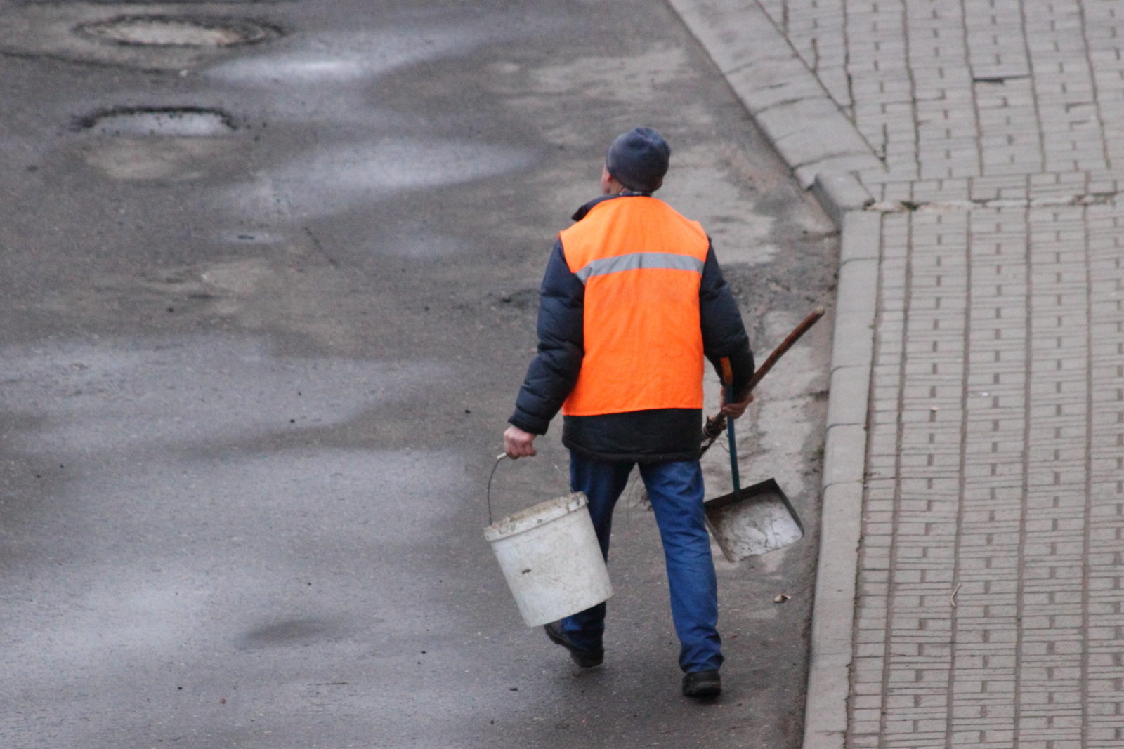 A janitor man in an orange vest walks with a bucket and scoop along an asphalt street, top view from behind