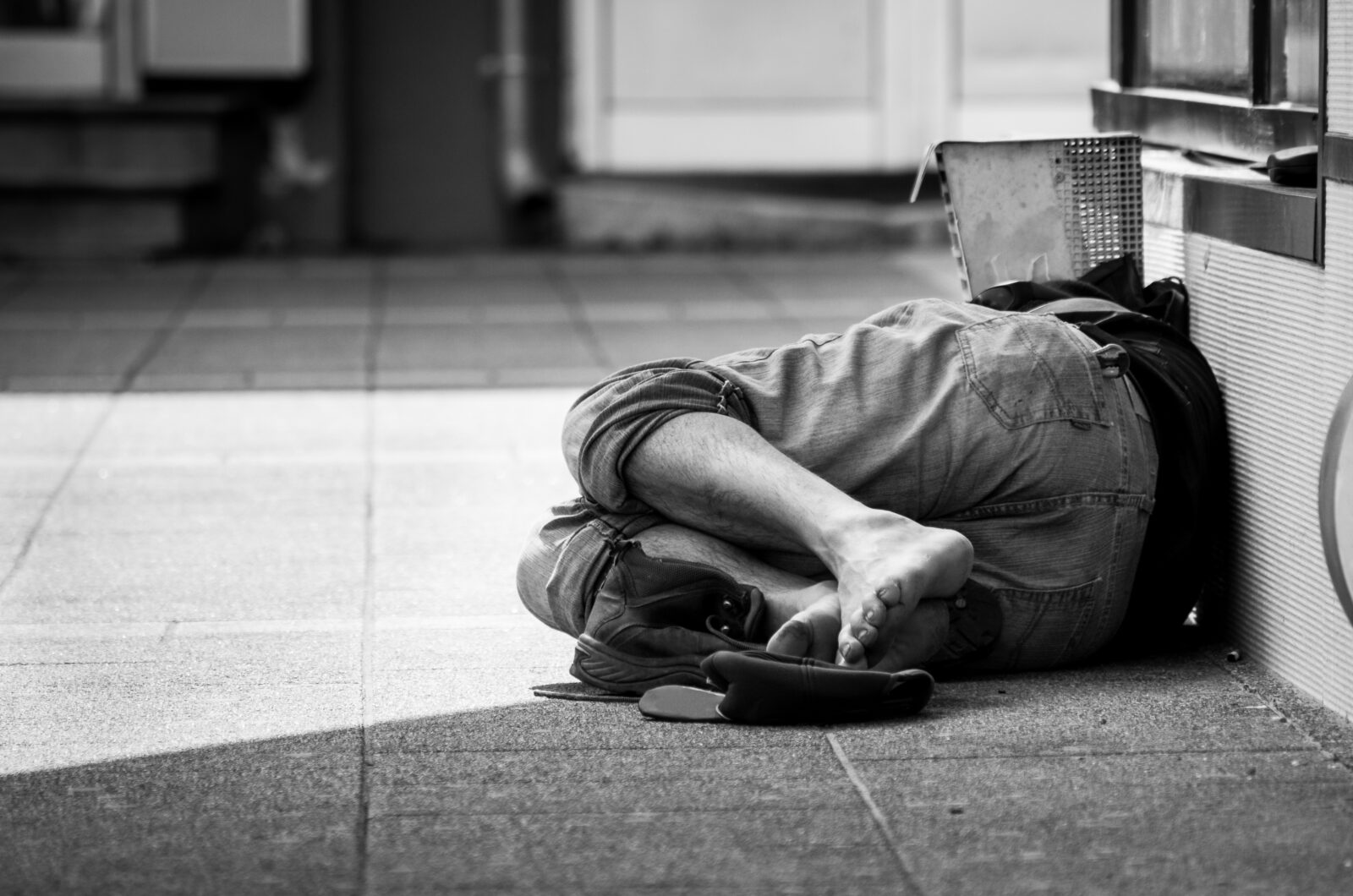 Poor homeless or refugee barefoot man sleeps on the street in the shadow of the building