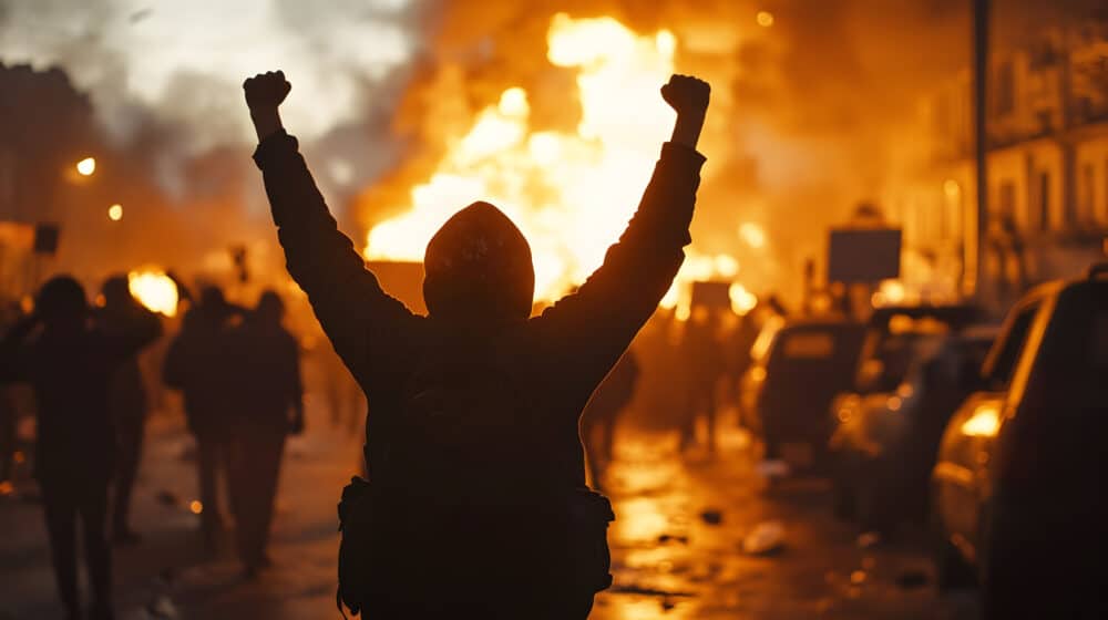 Intense urban protest at dusk with masked figure and fire in cityscape